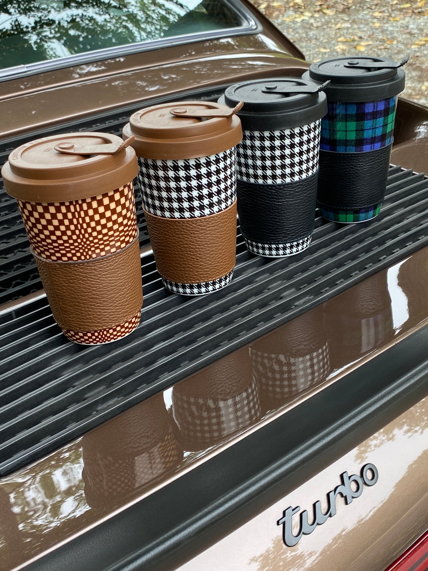 A porcelain "to-go" coffee cup featuring the Porsche Pepita Sport Classic pattern and a lid with a leather band in black, white, and brown.